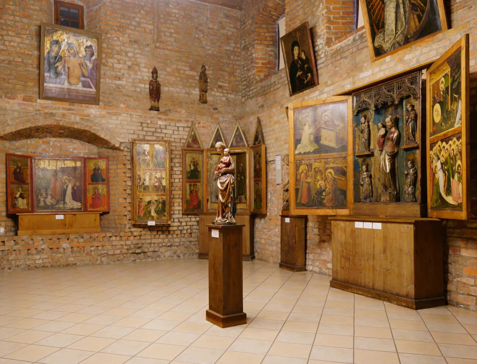 A museum hall with brick walls and tiled floors displays various religious artworks including painted panels and carved wooden statues, with a prominent statue of Mary holding baby Jesus on a pedestal in the center.