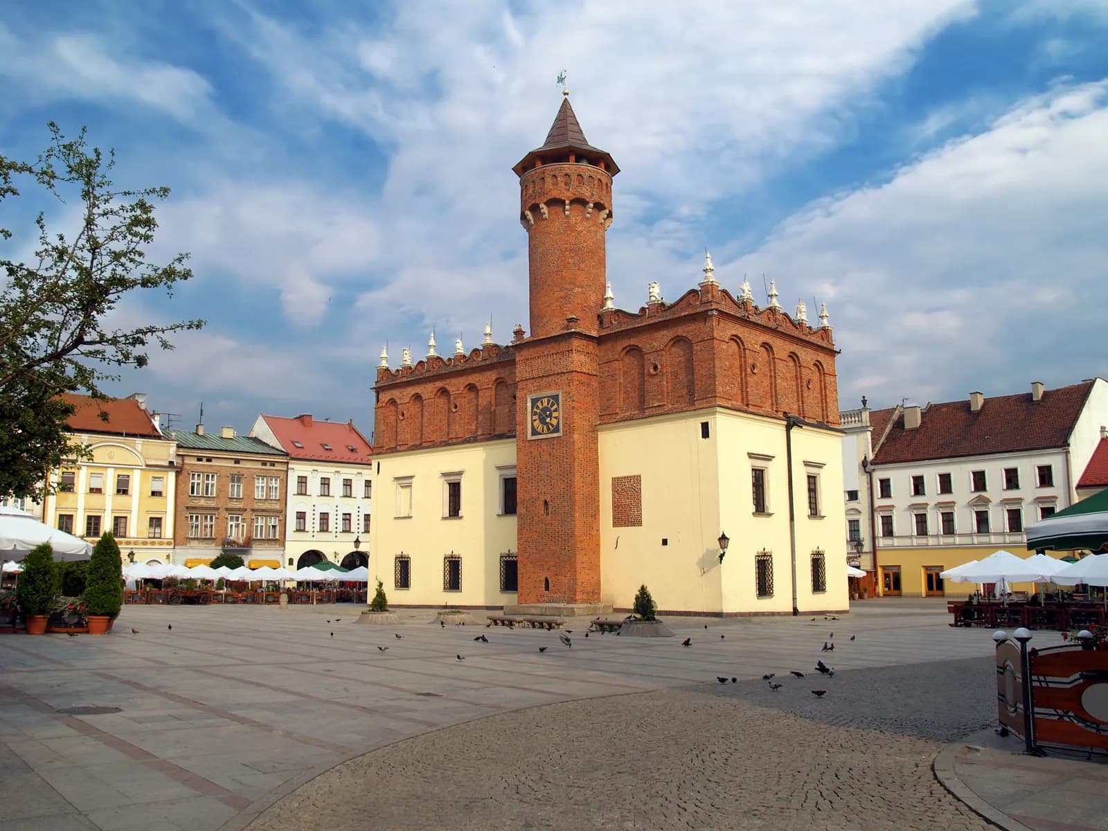 A large European town square features a dominant red brick tower with a clock and a cream-colored base, surrounded by other historic buildings and outdoor seating.